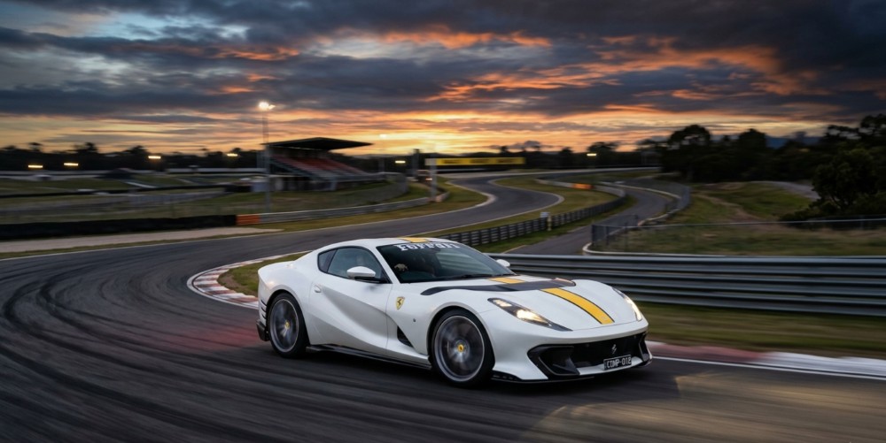 A white Ferrari 812 Competizione on a race track at dusk, motion blur on the rear wheels suggesting high speed.